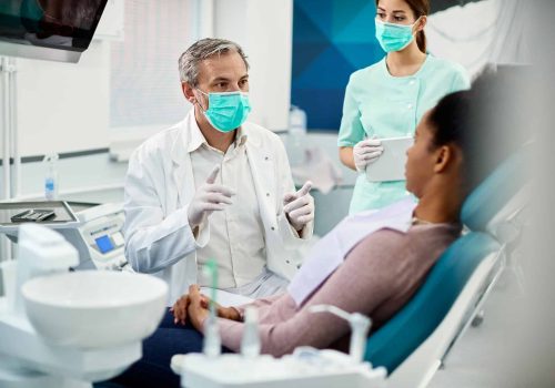 Mature dentist with face mask talking to black female patient at dentist's office.